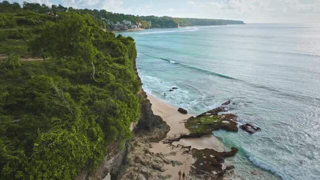 Aerial drone footage, flight over stunning coastline of Bali's Bukit Peninsula. Featuring lush green cliffs overlooking light sand beaches and clear turquoise ocean waves gently breaking on the shore