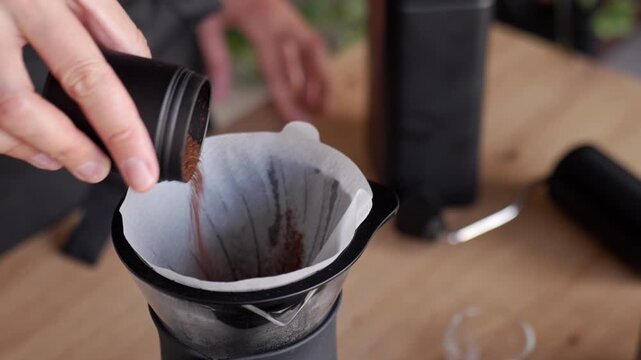 Person adding coffee grounds to paper filter before brewing pour over coffee hand pours ground coffee from a manual grinder into a dripper on a wooden table