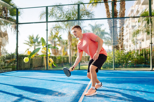 A young man on a blue tennis court holding a racket and ball. A man with a racket and ball practices on a paddle tennis court. Conceptual images of sports, activity, and weekends.