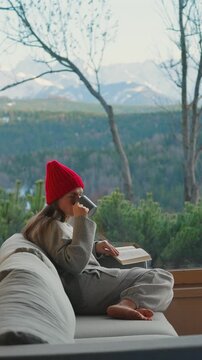 A woman reading by a large window and enjoying scenic snowy mountain view.