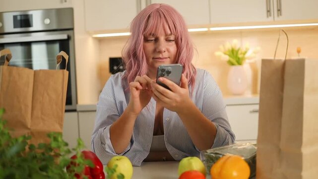 close-up shot of an attractive young woman with pink hair sitting in a bright kitchen, typing on her mobile phone near fresh vegetables and paper bags.