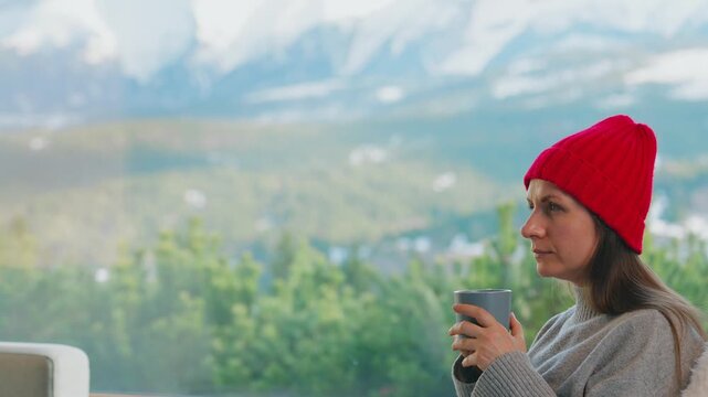 A woman sitting by a large window and enjoying scenic snowy mountain view.