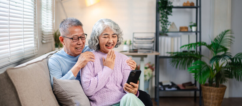 Happy Asian Older Couple Using Phone Sitting on Sofa at Home Smiling and Laughing, Looking at Screen Together, Enjoying Online Content and Having Fun