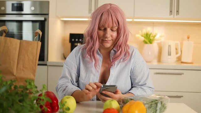 close-up shot of an attractive young woman with pink hair sitting in a bright kitchen, typing on her mobile phone near fresh vegetables and paper bags.