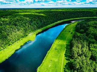 Obraz premium Flowing river meandering through dense green forest under a bright blue sky with clouds