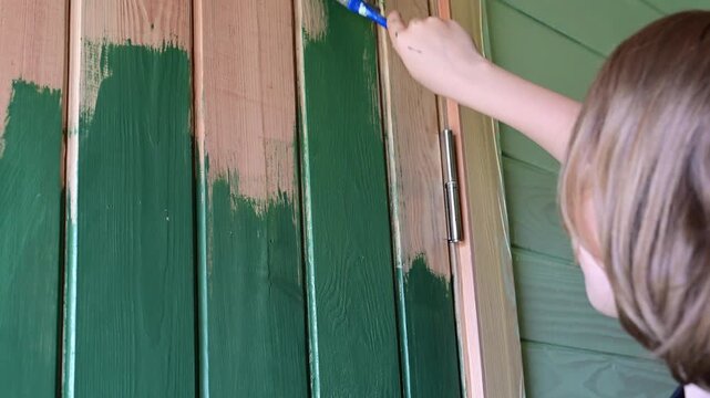 a child using a blue paintbrush to apply dark green paint to a vertically paneled wooden door.