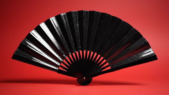 A sleek black handheld folding fan sits gracefully against a vibrant red background, subtly shifting its angle to catch the light in this minimalist and bold studio display.