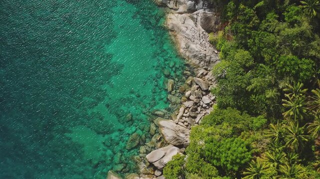 Phuket island coastline from above showing crystal clear turquoise water beside rocky granite shore and lush green tropical jungle with palm trees covering the landscape. Aerial top down background