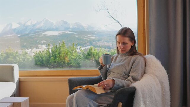 A woman reading by a large window and enjoying scenic snowy mountain view.