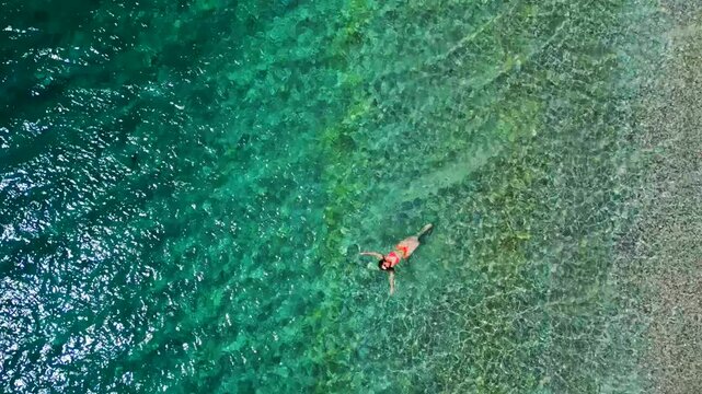 Woman in a red swimsuit swims on her back in the clear sea, top down view