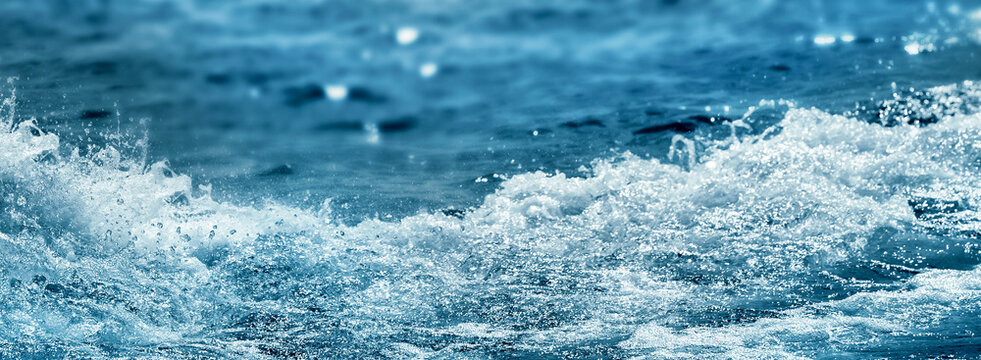 close-up of real splashing water wave with foam in foreground of blurred blue sea background, sunny fresh concept with copy space for travel destination, vacation, beach holiday, spa and body care