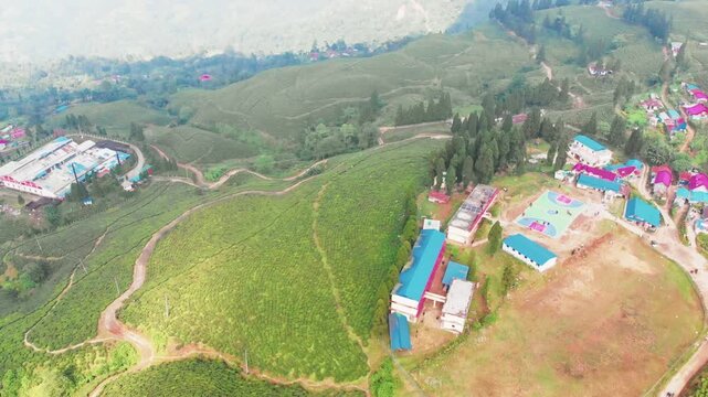 Wide aerial shot of Kanyam chiyabaari with a broad mountain highway running through tea-covered hills; village with basketball court visible , distant mountain ranges in the background
