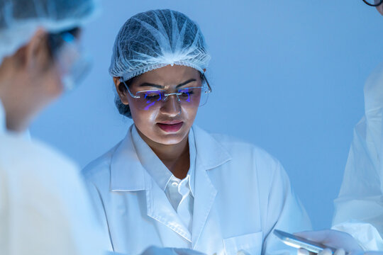 Portrait of woman in hairnet with digital reflections on eyeglasses looking down at work, colleagues nearby, high tech production concept.