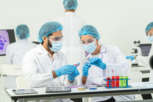 Man and woman in masks and hairnets examining red microscope slides together at table with test tube rack and tablets, medical research team.