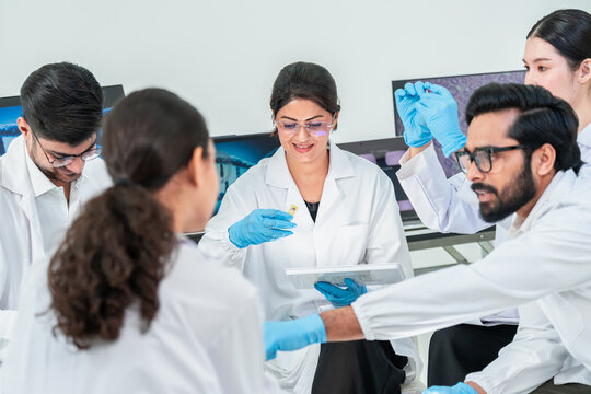 Smiling woman in glasses holding small specimen over tablet while colleagues gather around, collaborative laboratory team meeting.