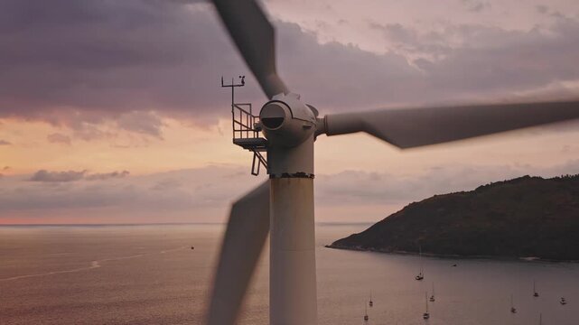 Close up wind turbine spinning its blades, generating clean sustainable electricity against a beautiful pink soft light sunset sky over the Andaman Sea, yachts and a tropical mountain on Phuket island