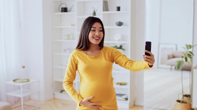 Smiling Asian Female In Her 20s, Expecting A Baby, Capturing Selfie While Standing In Bright Living Room At Home. Celebrating Motherhood And Enjoying Special Moments Of Pregnancy.