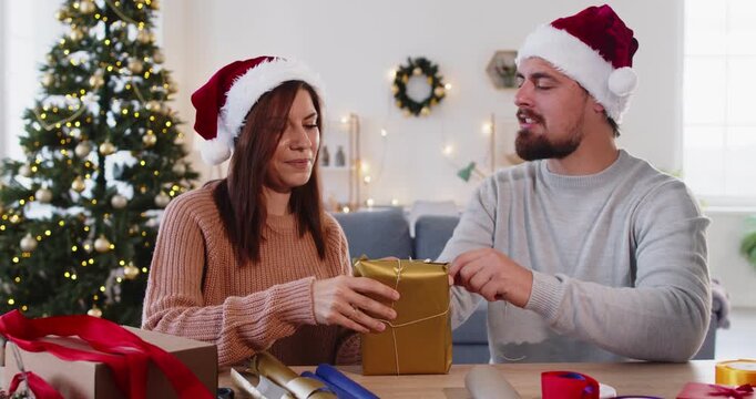 Christmas couple in Santa hats wrapping gift box with gold paper and ribbon at home. Happy man and woman tie thread and secure present with tape, people sitting at table with diy tools and materials