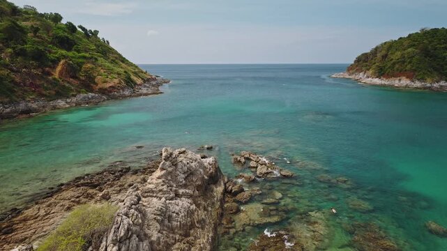 Phuket island's tropical coastline bay, lush green hills, rugged rock formations, and clear turquoise ocean water with people snorkeling in shallow reef enjoying the tranquil vacation. Drone flight