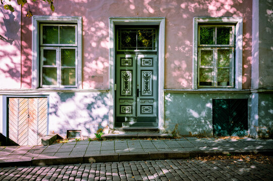Historical pink building facade in Tallinn with ornate wooden double door. Beautiful dappled sunlight shadows on street pavement. Old european city architecture