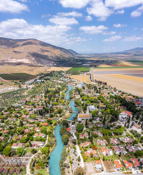 Aerial view of the kibbutz Nir David with its turquoise Asi stream flowing through lush greenery, surrounded by agricultural fields and mountains in Nir David, North District, Israel.