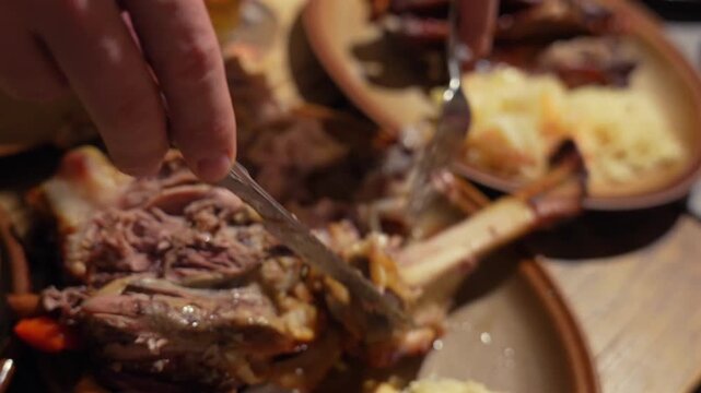 A hand carves Czech pork knuckle off the bone on a rustic ceramic plate with horseradish and pickled vegetables.