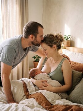Loving father kissing mother's forehead while breastfeeding their newborn baby in bedroom