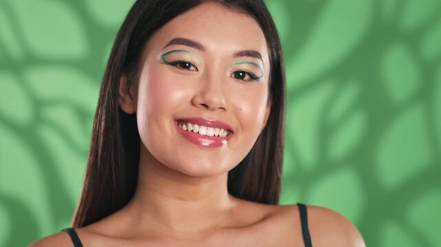 Smiling Asian Female Model With Bold Makeup Posing Confidently In Front Of Green Background. Capturing Beauty And Vibrancy At Studio. Adoration For Artistry And Self-expression. Beauty Concept.