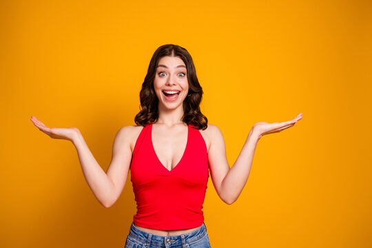 Happy young woman smiling and presenting with open palms against vibrant yellow background wearing red top and casual jeans
