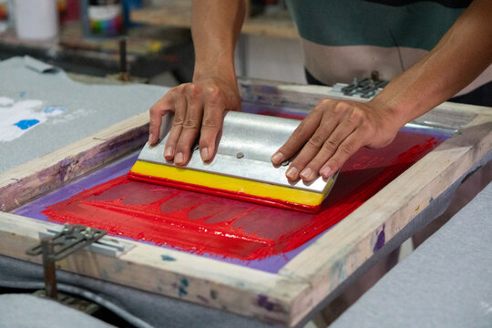 Hand uses a squeegee with a yellow rubber blade to push red ink through a mesh screen. This is a common manual screen printing process used to produce T-shirts.