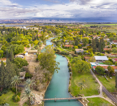 Aerial view of the Asi River flowing through lush greenery and residential areas, with a pedestrian bridge crossing the turquoise water in Nir David, North District, Israel.
