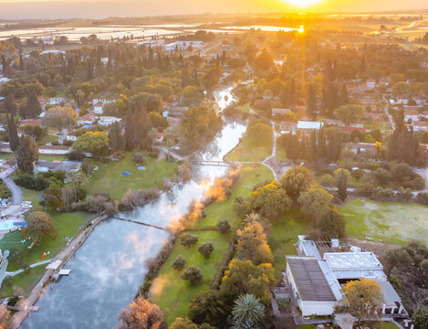 Aerial view of the Asi River flowing through the kibbutz at sunrise, with lush greenery, trees, and buildings under golden light in Nir David, North District, Israel.
