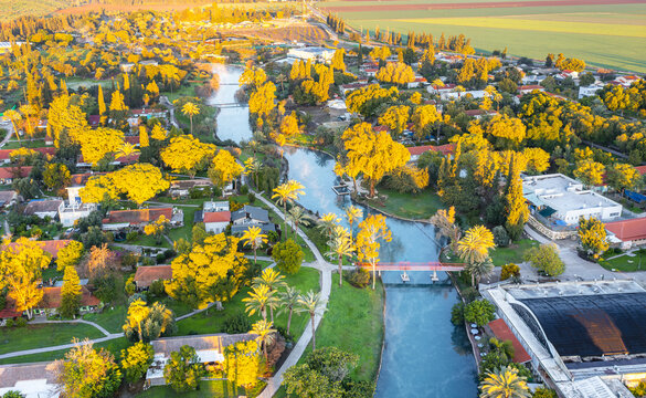 Aerial view of the kibbutz Nir David with its lush green trees, winding river, and red-roofed houses bathed in golden hour light in Nir David, North District, Israel.