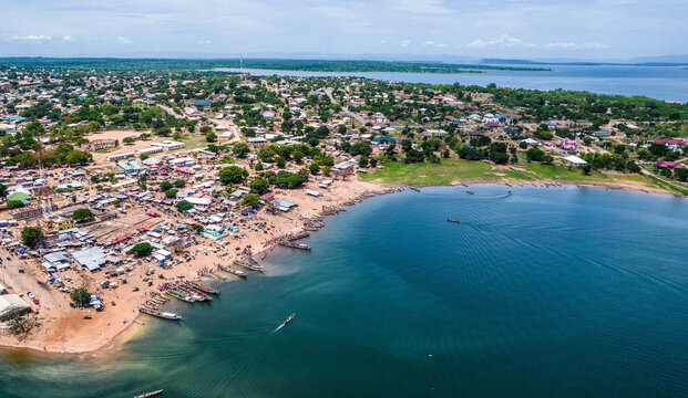 Aerial view of a bustling lakeside town with numerous small wooden fishing boats moored along the sandy shoreline, surrounded by residential houses and lush greenery in Ghana.