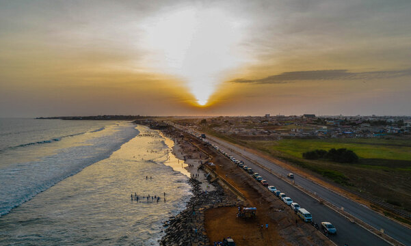 Aerial view of a coastal road at sunset with cars parked along the shoulder, people walking on the sandy beach, and waves rolling onto the shore in Ghana.