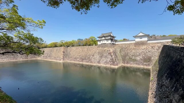 Historic Osaka Castle rises above its stone walls and moat beneath a clear blue sky in Osaka, Japan. A scenic travel destination framed by trees in early spring.
