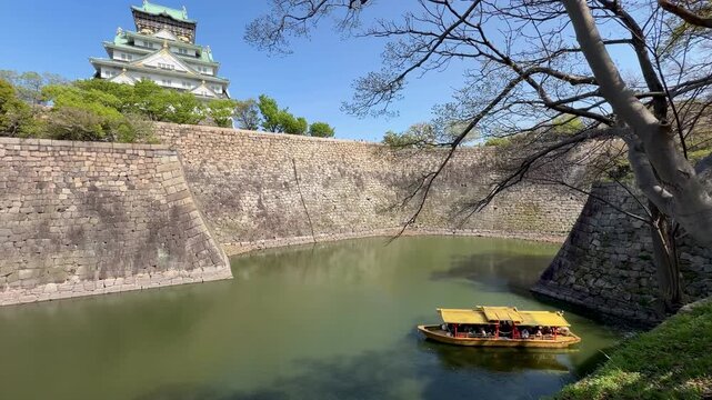 Historic Osaka Castle rises above its stone walls and moat beneath a clear blue sky in Osaka, Japan. A scenic travel destination framed by trees in early spring.