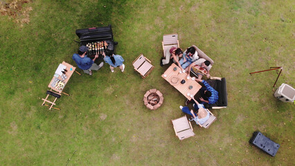 Fototapeta premium Aerial top-down view of a group of friends enjoying an outdoor barbecue party and camping on a green lawn, featuring a charcoal grill, wooden dining table, chairs, and a fire pit