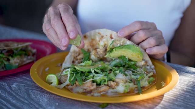 A senior woman squeezes lime on fish and shrimp tacos at a taco stand in Baja California. She smiles while enjoying her meal.