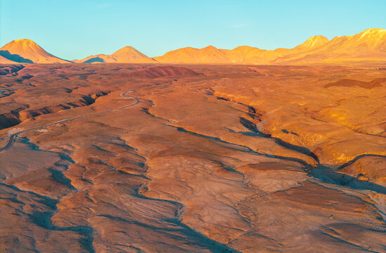 Aerial view of the arid desert landscape with rugged terrain, a winding dirt road, and distant volcanic mountains illuminated by golden hour light in Toconao, Antofagasta, Chile.