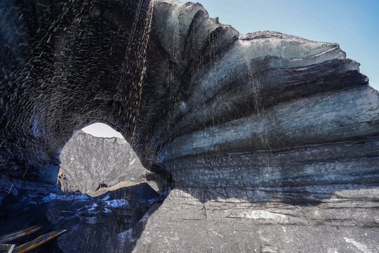 A natural ice arch formation with water cascading from melting glacier layers under a clear sky,Katla Ice Cave,Iceland