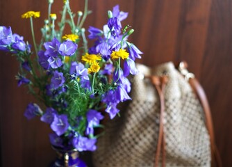 Summer bouquet of blue bells and yellow daisies in a blue vase on the background of a summer wicker straw bag on a wooden background © Irina