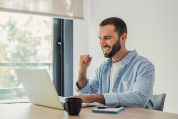 Fototapeta premium Happy bearded man celebrating a work victory, raising fist in air while sitting at desk in a home office, achieving success online with his computer