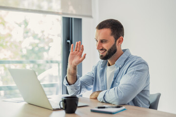 Naklejka premium Young man smiling and waving to a laptop screen during a video call, communicating remotely from his modern home office setup while engaging in virtual conversations