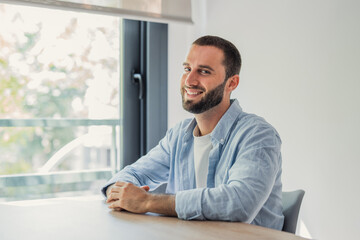 Fototapeta premium Smiling young man in a light blue shirt sits at a home office desk near a sunny window, conveying relaxed confidence, remote work lifestyle, productivity and approachable professionalism.