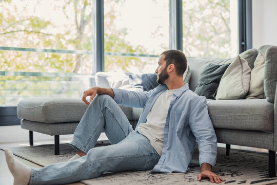 Young man sitting on the living room floor leaning against a gray sofa, looking through a large window with green trees outside, feeling reflective and calm at home