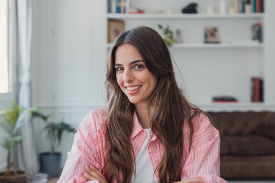 Young happy woman smiling confidently at the camera, posing with crossed arms in a comfortable indoor setting, representing well-being, positivity, and a relaxed lifestyle