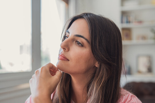 Young brunette woman lost in thought, looking away, reflecting on ideas and dreams indoors with natural window light highlighting her face