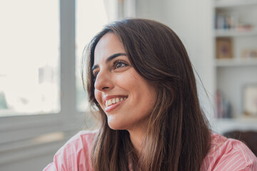 Smiling young woman looks out a bright window in a relaxed, casual home setting. Natural light highlights her thoughtful, happy expression and warm, approachable mood in an intimate portrait. © Daniel