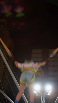 Child leaping high into the night air on a bungee trampoline at an amusement park, held by stretched elastic cords and a harness, floodlights and apartment towers below